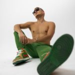 A shirtless man poses in green pants and shoes.